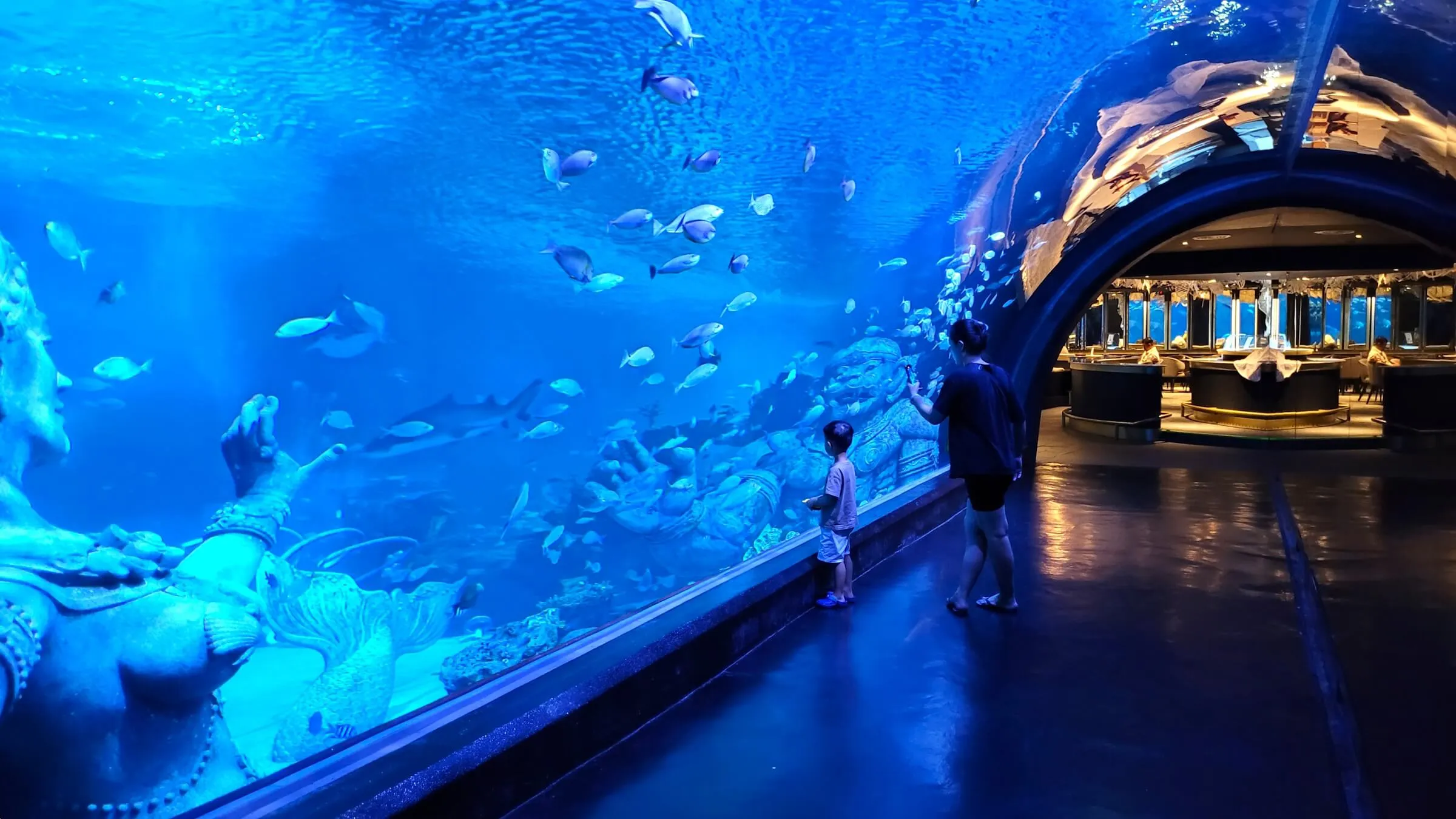 A child and a woman at the long acrylic wall of the main tank at Aquaria Phuket, with fish, a shark and Thai-style statues behind the glass